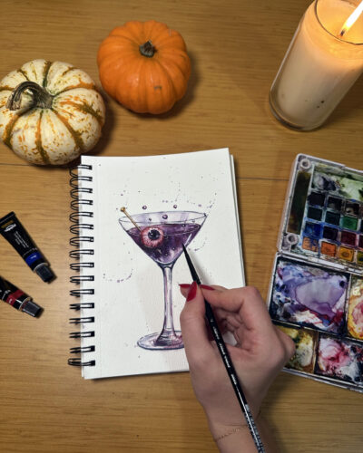 Artist painting a spooky purple martini with an eyeball garnish during a Halloween watercolor sip and paint workshop by Tamila Dubas and LaLaLu NYC at Bird in Hand, Hamilton Heights.